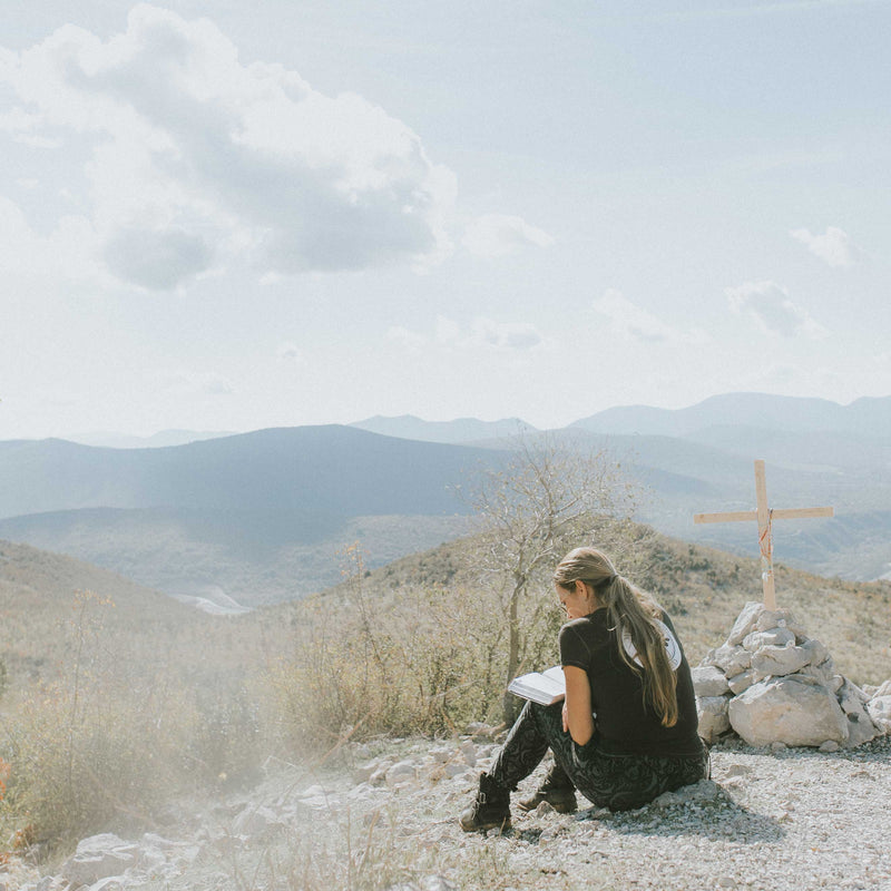 Woman seated on ground next to wooden cross reading, overlooking canyons.