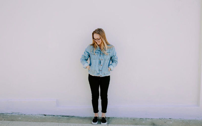young woman wearing Divine Mercy Blessing bracelets, denim jacket and black pants standing against a plain outdoor wall hands in pockets