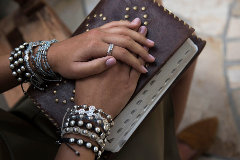 young woman's hands folded on bible, wearing Hail Mary Morse Code bracelet and other My Saint My Hero Blessing Bracelets