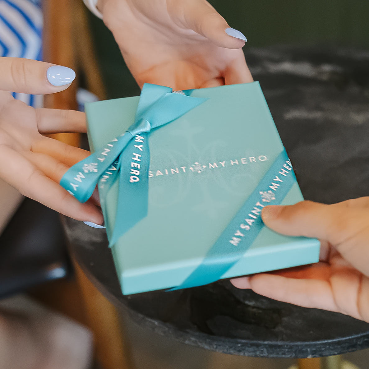 Two hands holding a turquoise gift box with 'My Saint My Hero' branding.
