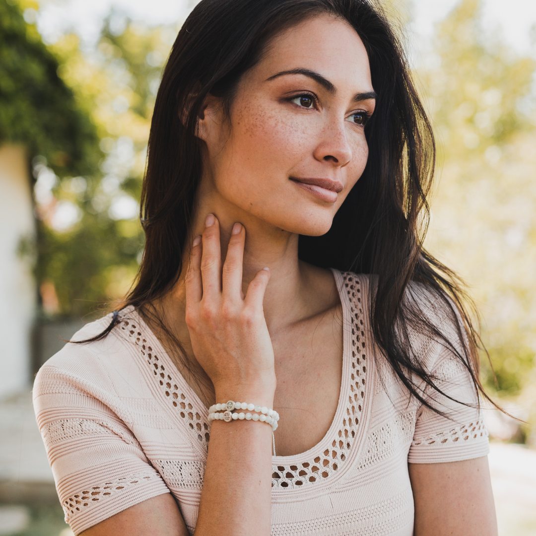 woman with her back to camera, staring out at the beach ocean, wearing tan sheer top with her arms over her head and shoulders, each arm has three or four black silver and gold woven saint medal bracelets