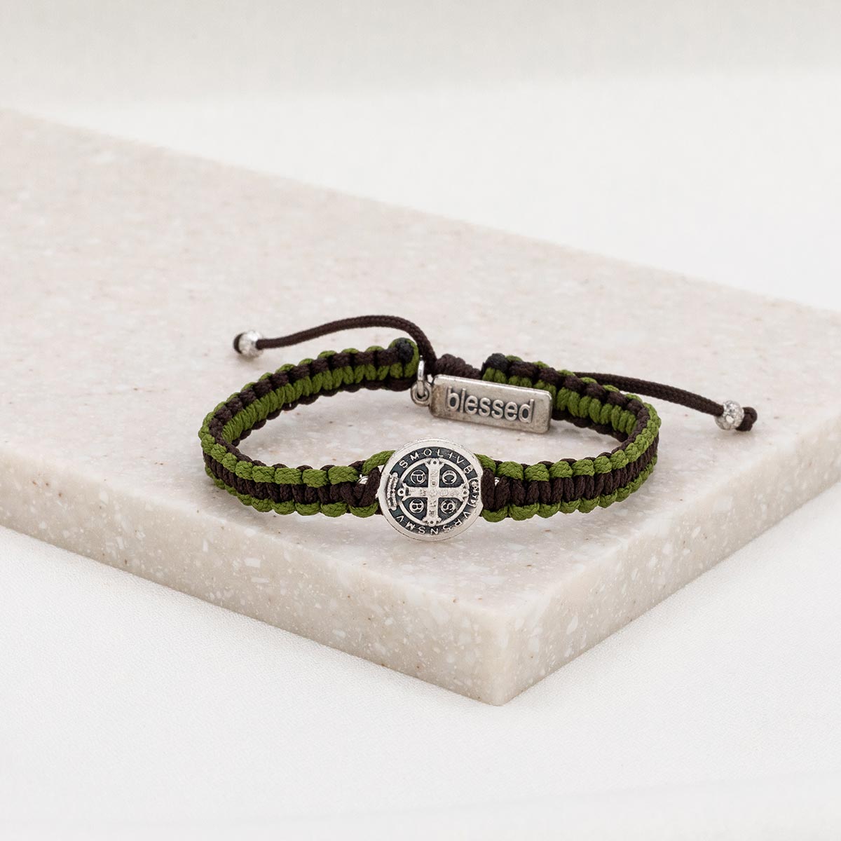 A green and brown woven bracelet with a silver-tone Benedictine medal and a silver-tone 'blessed' tag, displayed on a marble surface.
