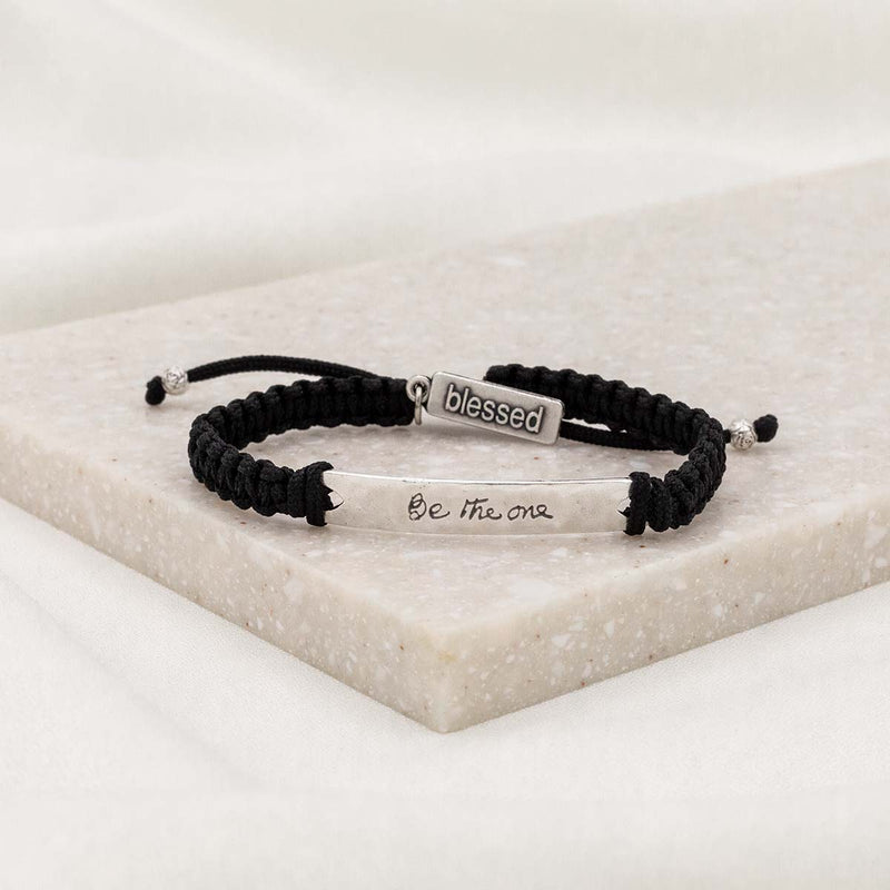 A black adjustable bracelet with a silver-tone hammered zinc bar, engraved with the words 'Be the one' and the signature 'blessed' tag, displayed on a white marble surface.