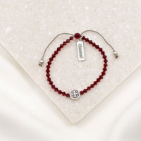 A beaded bracelet with deep red faceted beads, a silver adjustable cord, a round silver charm, and a small rectangular silver tag engraved with 'obessed'. The bracelet is displayed on a textured cream-colored stone tile.