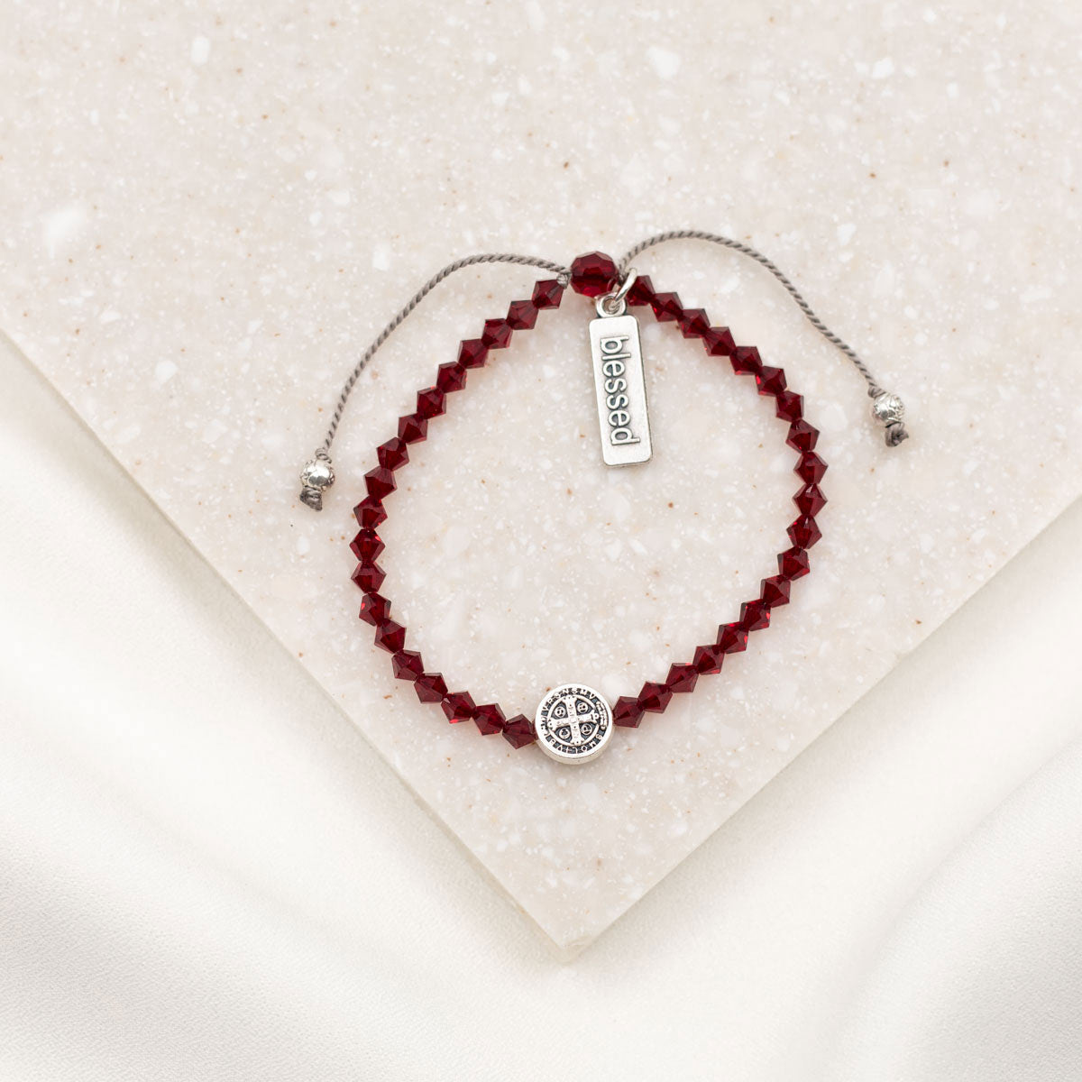 A beaded bracelet with deep red faceted beads, a silver adjustable cord, a round silver charm, and a small rectangular silver tag engraved with 'obessed'. The bracelet is displayed on a textured cream-colored stone tile.