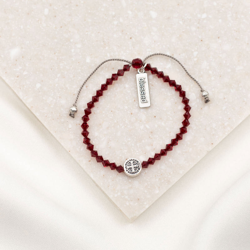 A beaded bracelet with deep red faceted beads, a silver adjustable cord, a round silver charm, and a small rectangular silver tag engraved with 'obessed'. The bracelet is displayed on a textured cream-colored stone tile.