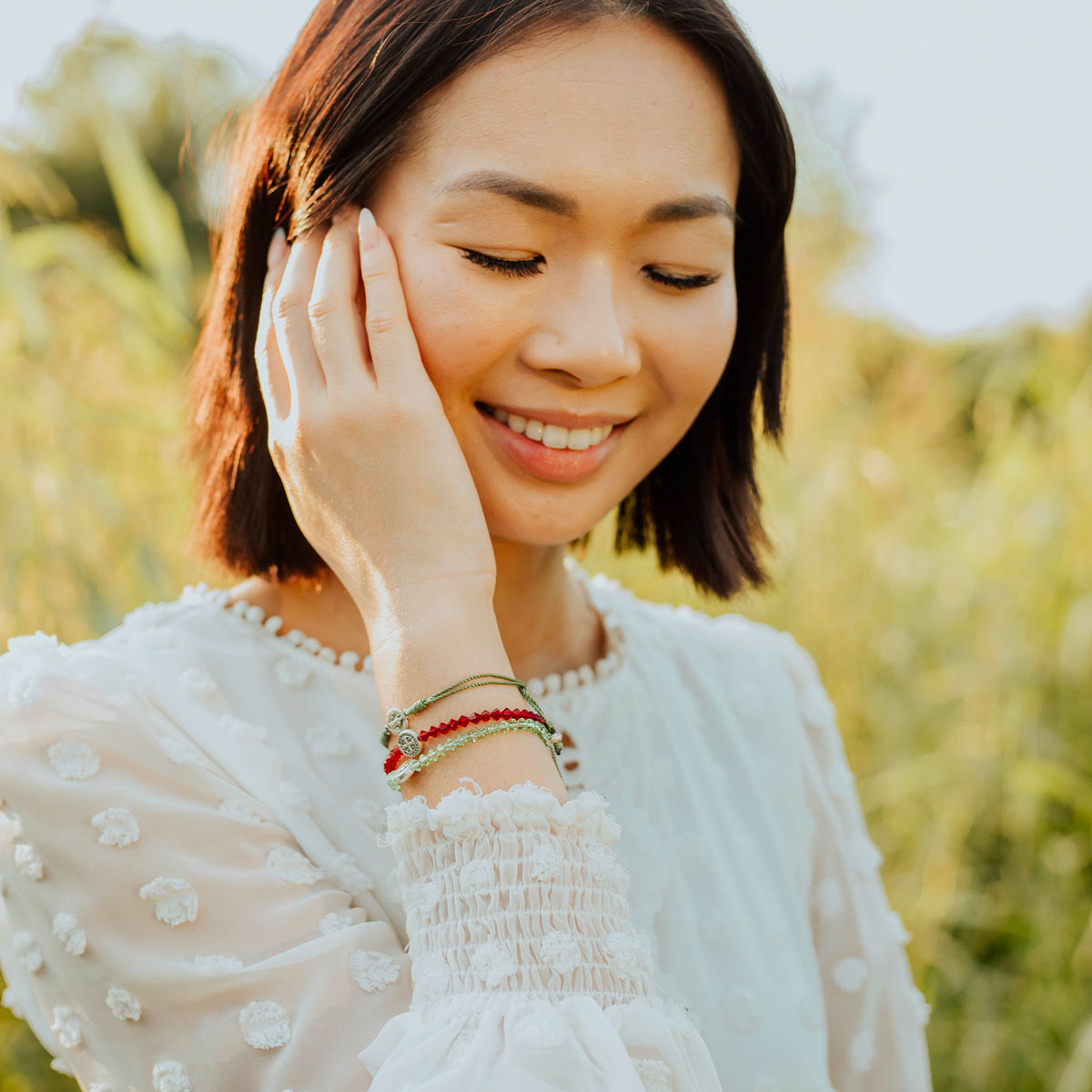 A woman with short brown hair wearing a white textured blouse and several colorful beaded bracelets, posing outdoors with a soft-focus green background.