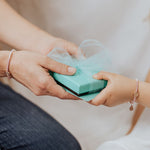 mother handing a gift box to daughter close up of hands both wearing love lights the way crystal pearl matching bracelets