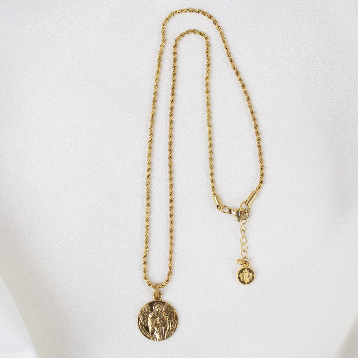 Gold round Archangel Gabriel guardian angel medallion pendant necklace displayed on its matching gold rope chain, showcasing the full length of the necklace, its adjustable lobster clasp closure, and a small branded "My Saint My Hero" logo charm, resting flat against a clean, white textured stone background.