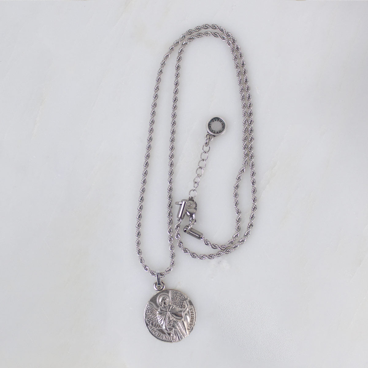 Silver round Archangel Gabriel guardian angel medallion pendant necklace displayed on its matching silver rope chain, showcasing the full length of the necklace and its adjustable lobster clasp closure, resting flat against a clean, white textured stone background.