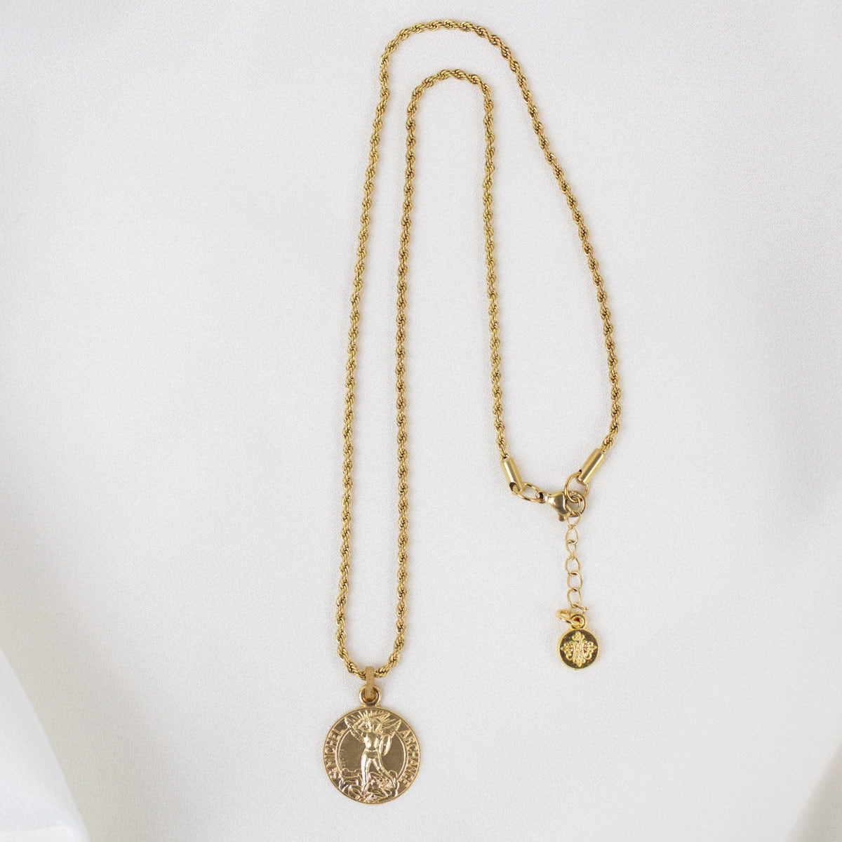 Gold round Archangel Michael guardian angel medallion pendant necklace displayed on its matching gold rope chain, showcasing the full length of the necklace, its adjustable lobster clasp closure, and a small branded "My Saint My Hero" logo charm, resting flat against a clean, white textured stone background.
