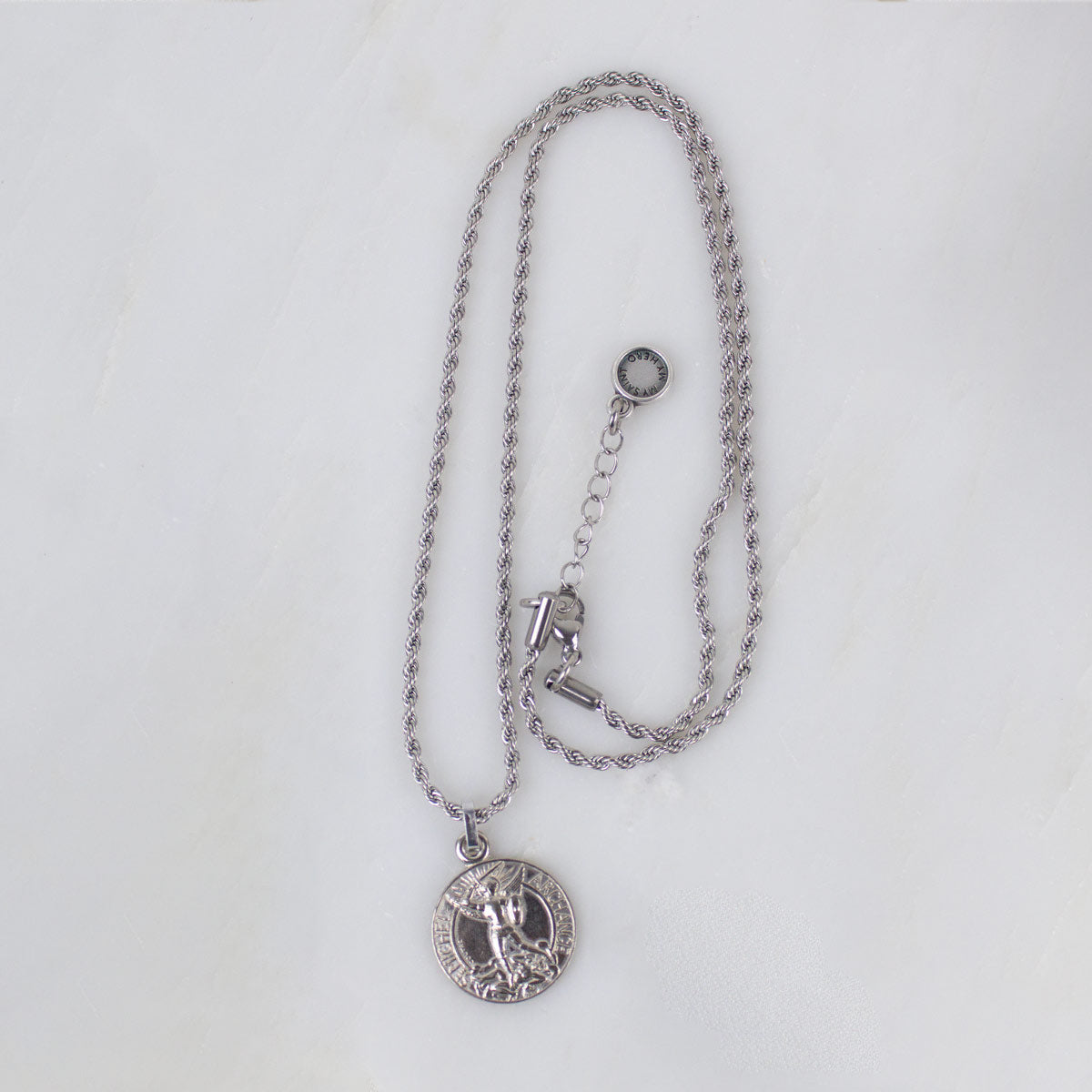 Silver round Archangel Michael guardian angel medallion pendant necklace displayed on its matching silver rope chain, showcasing the full length of the necklace and its adjustable lobster clasp closure, resting flat against a clean, white textured stone background.