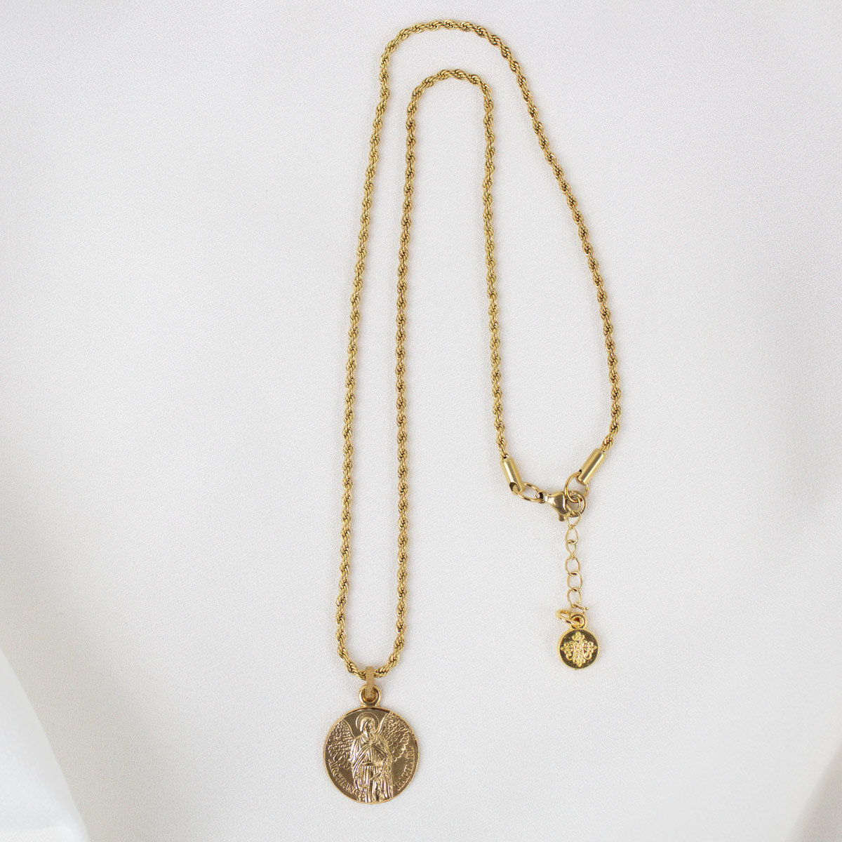 A full-length view of the gold Archangel Raphael Guardian Angel medallion necklace displayed against a smooth, white background. The round medal featuring the detailed engraving of the angel hangs from a long, delicate gold rope chain. The chain is arranged in a vertical loop, showing the clasp closure at the top right, complete with an adjustable extender chain and a small, round branded metal tag hanging at the end.