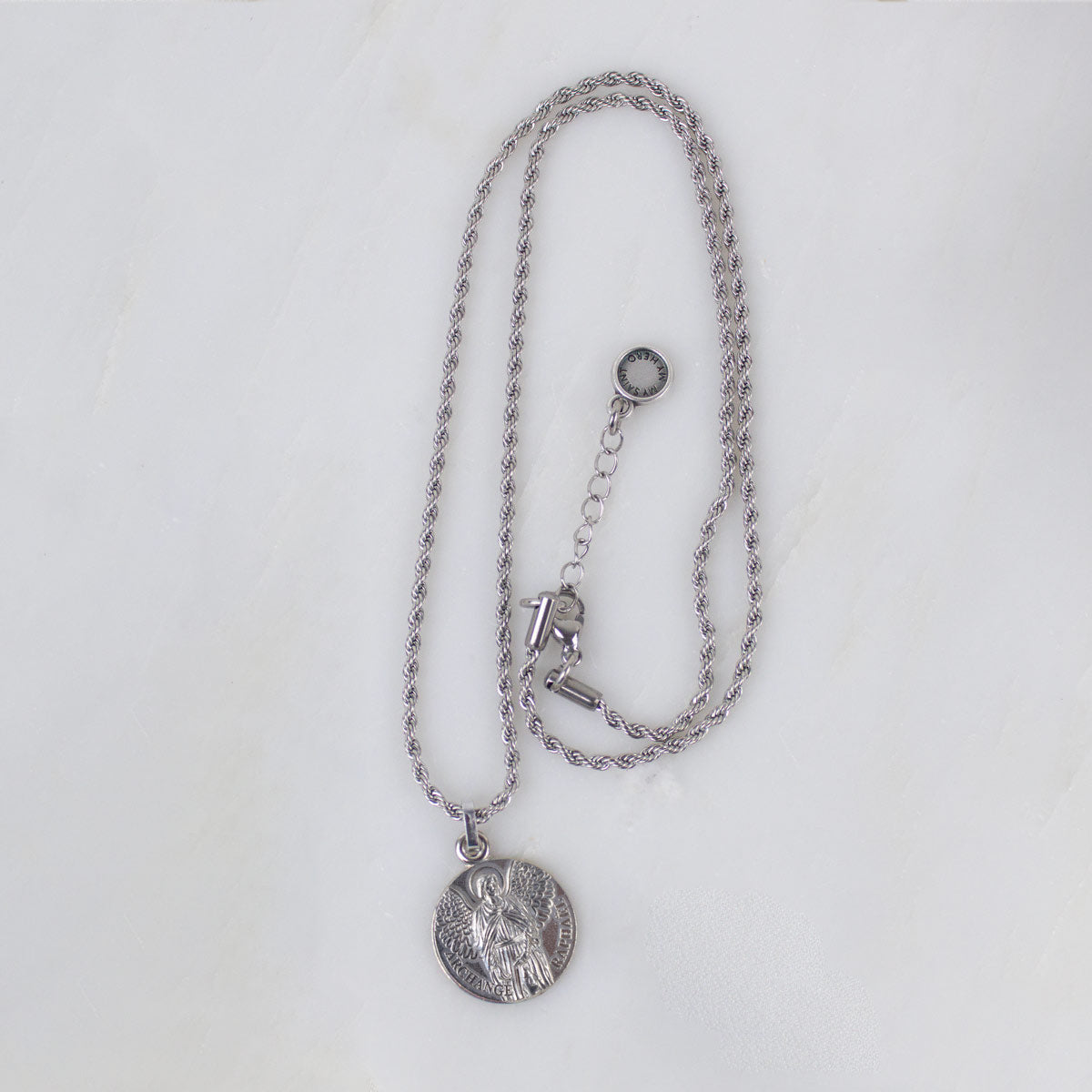 A gold Archangel Raphael Guardian Angel medallion necklace resting on a light-colored, textured stone surface. The round medal features a detailed engraving of the angel with wings spread, holding a staff. The medallion is attached to a matching gold rope-style chain.