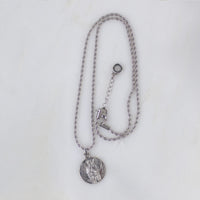 A gold Archangel Raphael Guardian Angel medallion necklace resting on a light-colored, textured stone surface. The round medal features a detailed engraving of the angel with wings spread, holding a staff. The medallion is attached to a matching gold rope-style chain.