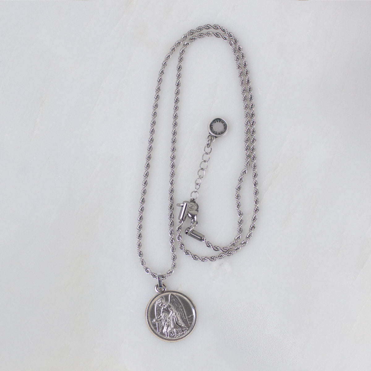 A top-down, full-length view of the silver Archangel Raphael Guardian Angel medallion necklace laid out on a clean white background. The round medal with its intricate engraving hangs from a long, delicate silver rope chain. The chain forms a vertical loop, leading up to a secure lobster-style clasp on the right side, an adjustable extender chain, and a tiny, round branded tag hanging at the very end.