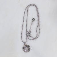 A top-down, full-length view of the silver Archangel Raphael Guardian Angel medallion necklace laid out on a clean white background. The round medal with its intricate engraving hangs from a long, delicate silver rope chain. The chain forms a vertical loop, leading up to a secure lobster-style clasp on the right side, an adjustable extender chain, and a tiny, round branded tag hanging at the very end.