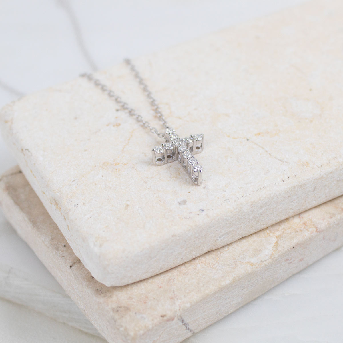 A silver cross pendant necklace with small clear stones, resting on stacked light beige stone blocks.