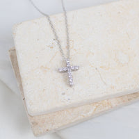 A silver cross pendant necklace with small clear stones, resting on a textured beige stone block.