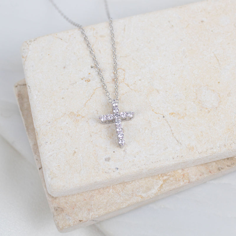 A silver cross pendant necklace with small clear stones, resting on a textured beige stone block.