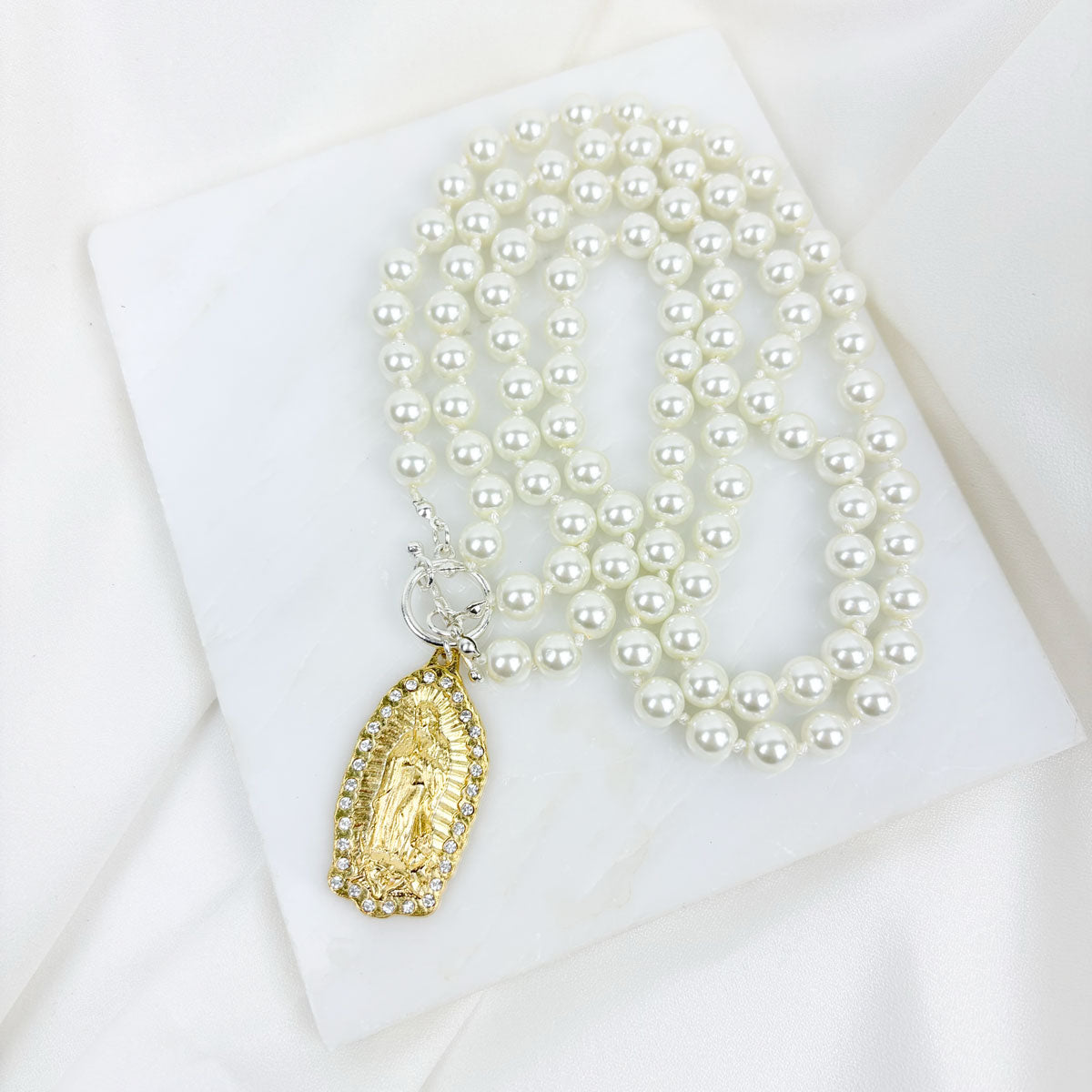 A pearl necklace with a silver-tone toggle and a gold-plated Our Lady of Guadalupe medal with hand-laid crystals, displayed on a white surface.