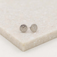 A pair of silver-tone stud  st. benedict medal earrings with embossed design, displayed on a beige surface.