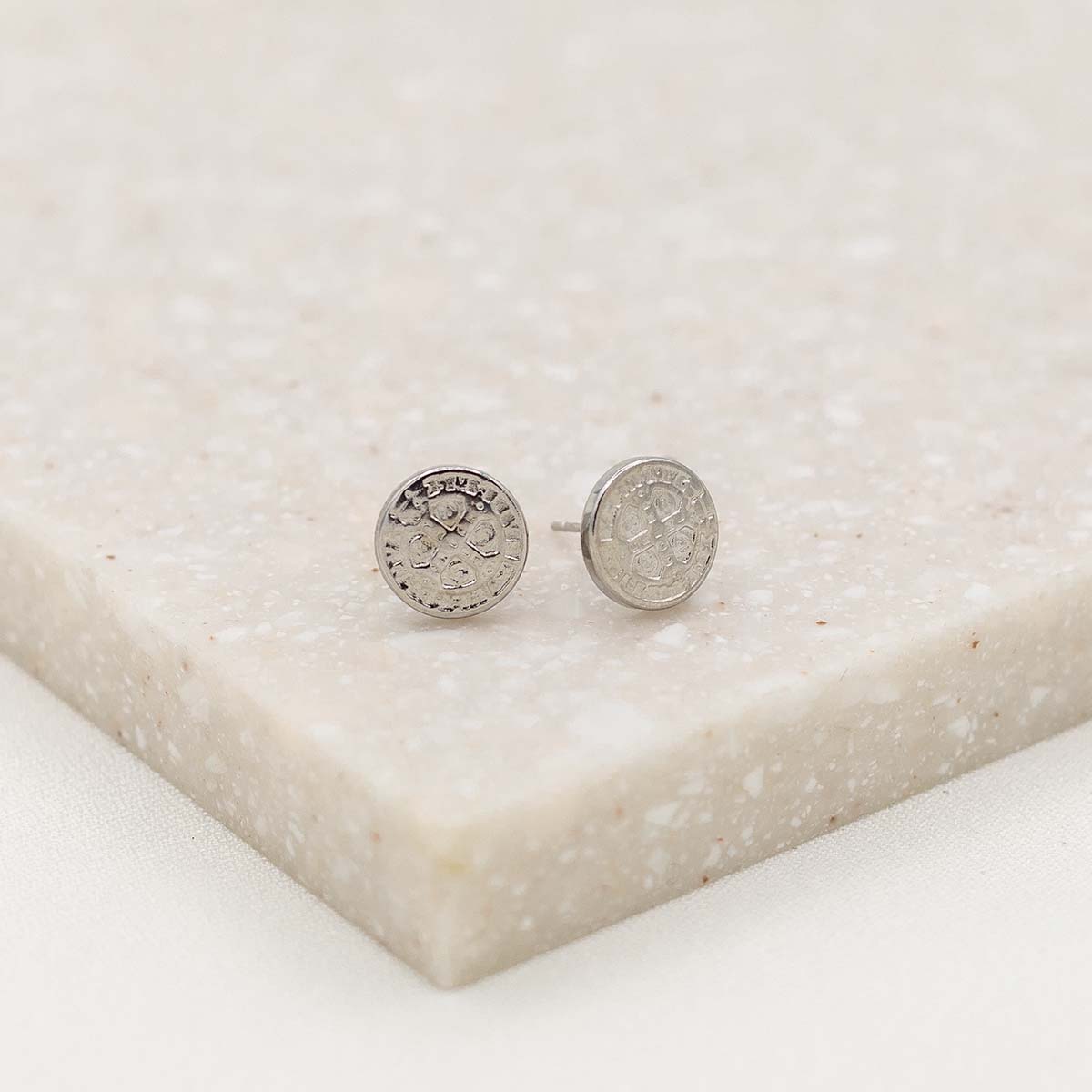 A pair of silver-tone stud  st. benedict medal earrings with embossed design, displayed on a beige surface.