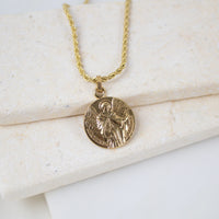 Gold round Archangel Gabriel guardian angel medallion pendant on a gold rope chain, resting on a white textured stone surface. The medal features a detailed engraving of St. Gabriel holding a lily and a scroll, with the inscription "S. GABRIEL ARCHANGE".