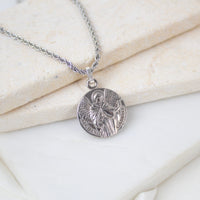 Silver round Archangel Gabriel guardian angel medallion pendant on a silver rope chain, resting on a white textured stone surface. The medal features a detailed engraving of St. Gabriel holding a lily and a scroll, with the inscription "S. GABRIEL ARCHANGE".