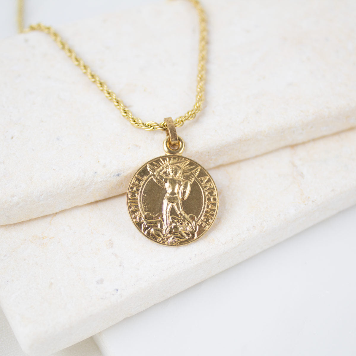  Gold round Archangel Michael guardian angel medallion pendant on a gold rope chain, resting on a white textured stone surface. The medal features a detailed engraving of St. Michael defeating a dragon with the inscription "S. MICHEL ARCHANGE".