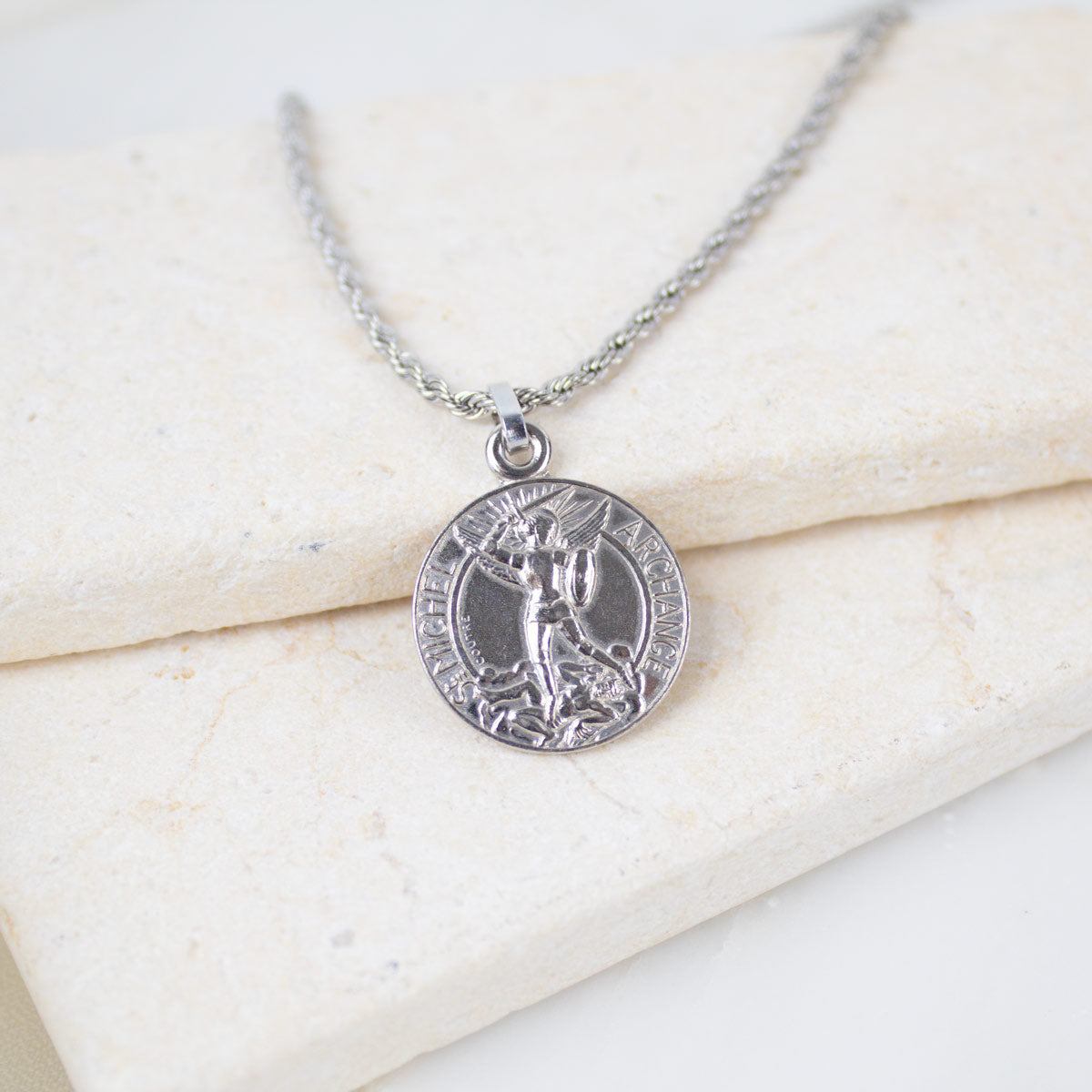 Silver round Archangel Michael guardian angel medallion pendant on a silver rope chain, resting on a white textured stone surface. The medal features a detailed engraving of St. Michael defeating a dragon with the inscription "S. MICHEL ARCHANGE".