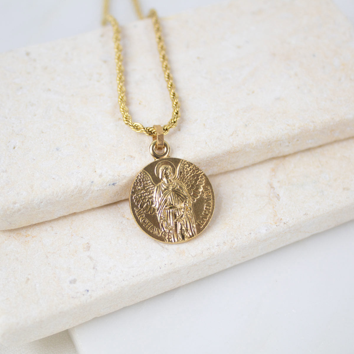 A gold Archangel Raphael Guardian Angel medallion necklace resting on a light-colored, textured stone surface. The round medal features a detailed engraving of the archangel with wings spread, holding a staff. The medallion hangs from a matching gold rope-style chain.