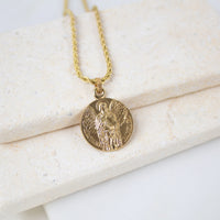 A gold Archangel Raphael Guardian Angel medallion necklace resting on a light-colored, textured stone surface. The round medal features a detailed engraving of the archangel with wings spread, holding a staff. The medallion hangs from a matching gold rope-style chain.