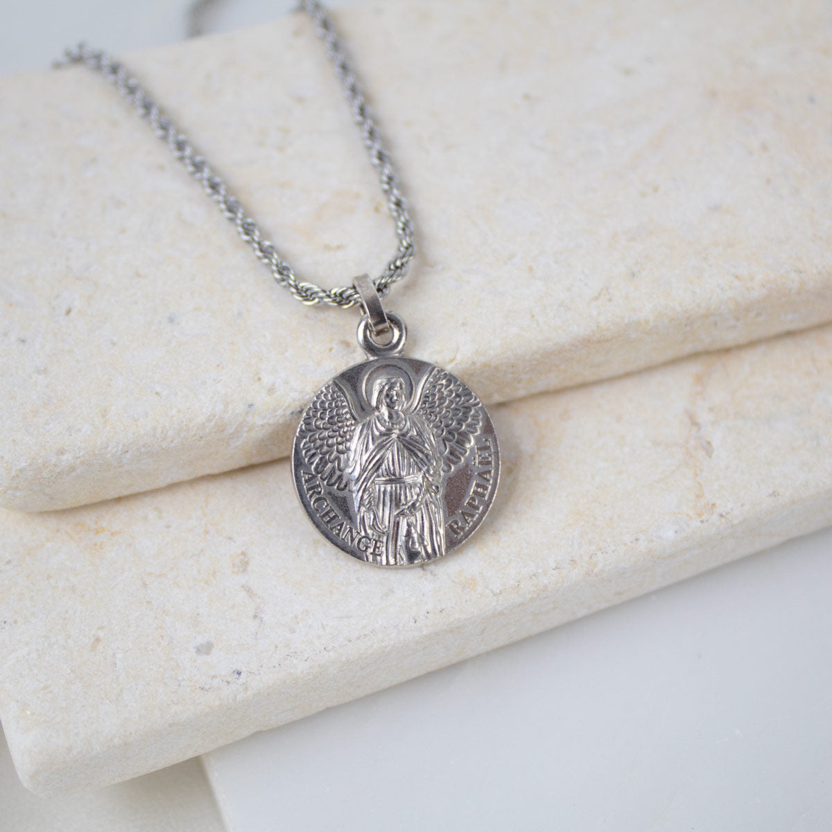 A close-up of a silver Archangel Raphael Guardian Angel medallion necklace. The round medal features a detailed engraving of the archangel with wings spread, holding a staff, with the words "ARCHANGEL RAPHAEL" inscribed along the bottom edge. The medallion hangs from a silver rope-style chain and rests on a light-colored, textured stone surface.