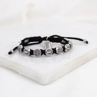 A black cord bracelet with silver-tone Benedictine Medals and a "blessed" charm tag.