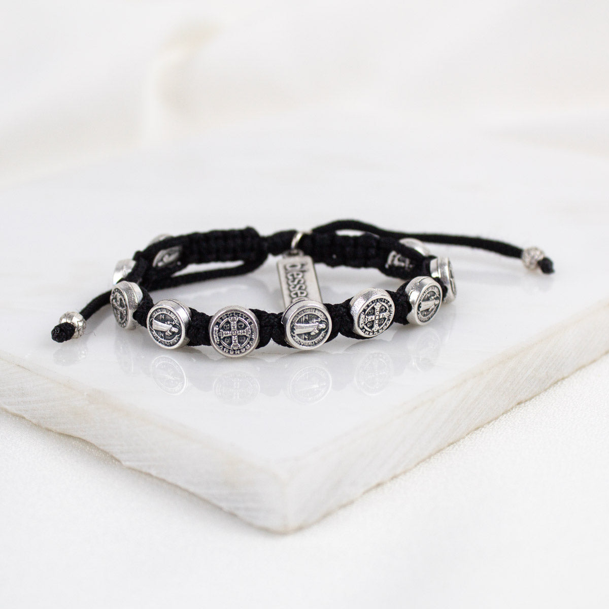 A black cord bracelet with silver-tone Benedictine Medals and a "blessed" charm tag.