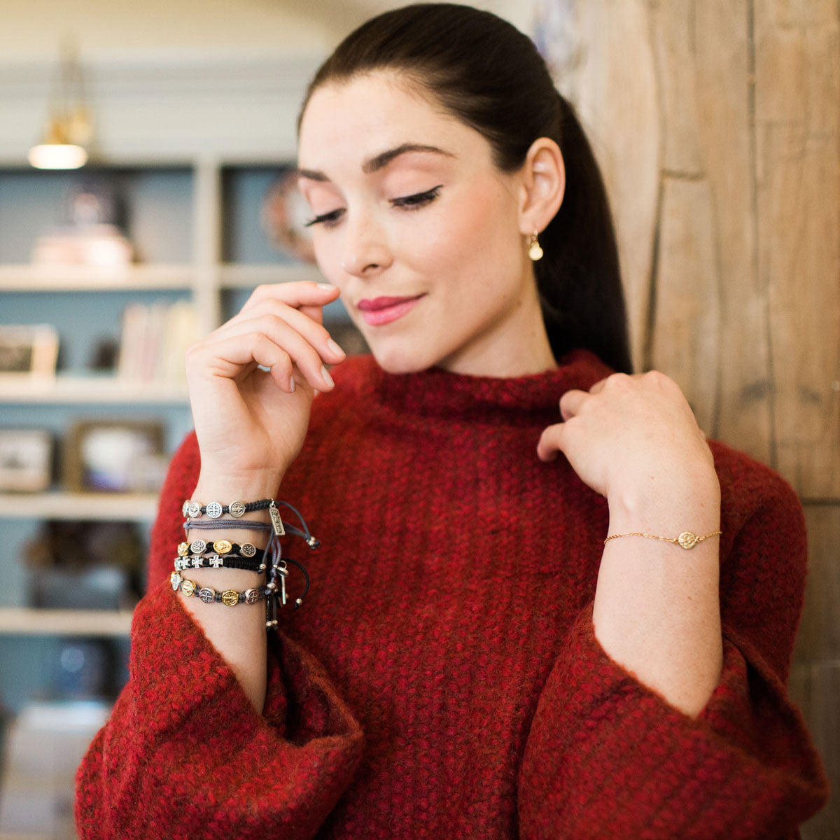 woman in red sweater wearing a gold breathe chain bracelet and woven my saint my hero bracelets