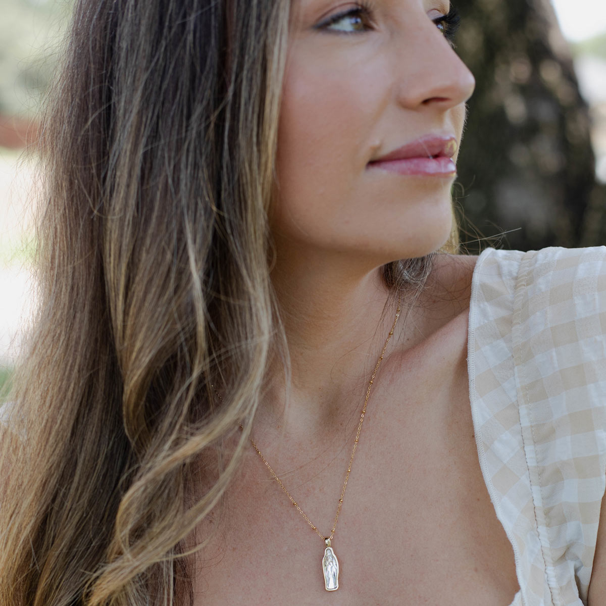 A close-up, side-profile view of a woman outdoors with long, wavy blonde hair, wearing a white and beige gingham checkered top. She is wearing a delicate gold station chain necklace featuring a petite, vertical, clear crystal rectangular pendant.