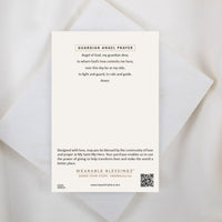 The back of an inspirational product card titled "GUARDIAN ANGEL PRAYER" displayed on a white marble surface. It features a traditional prayer that reads: “Angel of God, my guardian dear, to whom God’s love commits me here, ever this day be at my side, to light and guard, to rule and guide. Amen.” The bottom of the card includes a brief note about the "Wearable Blessings" community, a brand logo, and a QR code.