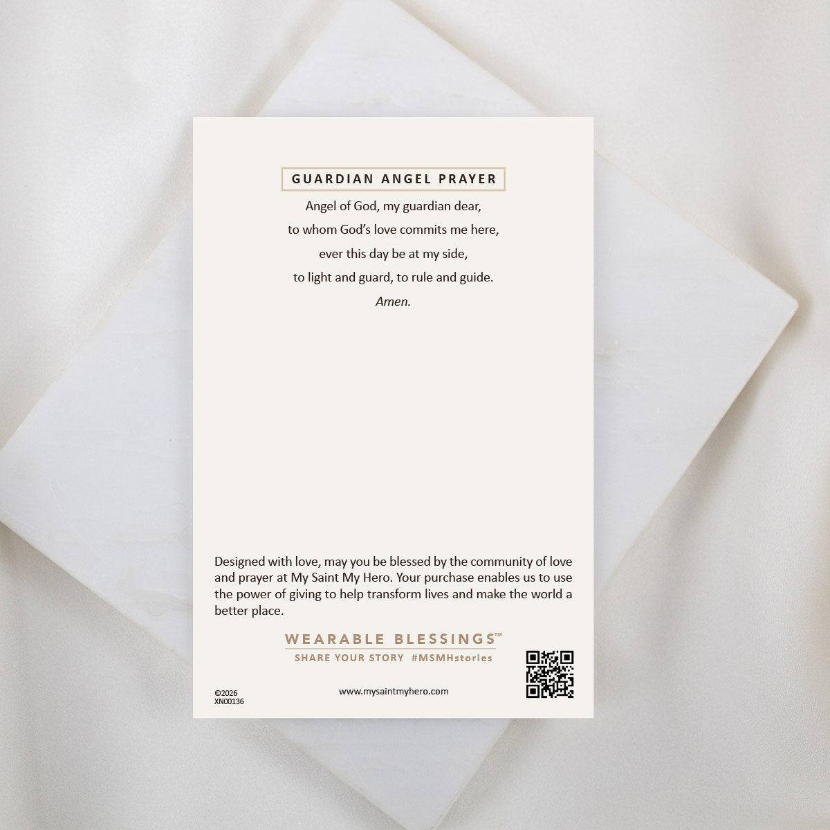The back of an inspirational product card titled "GUARDIAN ANGEL PRAYER" displayed on a white marble surface. It features a traditional prayer that reads: “Angel of God, my guardian dear, to whom God’s love commits me here, ever this day be at my side, to light and guard, to rule and guide. Amen.” The bottom of the card includes a brief note about the "Wearable Blessings" community, a brand logo, and a QR code.