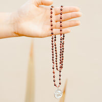 Hand holding a long beaded necklace with a pendant against a beige background