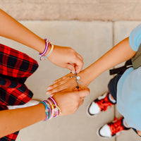a child wearing a stack of Pray. Hope. Don't Worry. Blessing Bracelets putting a bracelet with a happy face on another student's hand
