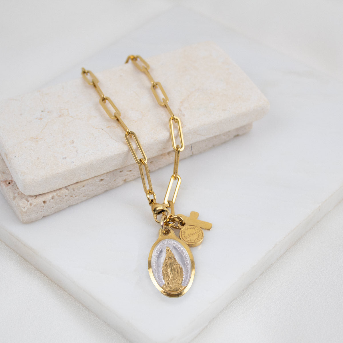 Gold paper clip bracelet with an Our Lady of Lourdes  pendant and a cross on a white surface
