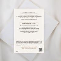 The back of a My Saint My Hero informational product card for the Archangel Gabriel Guardian Angel Necklace, resting on a white textured stone surface. The cream-colored card features the heading "ARCHANGEL GABRIEL" explaining his name and role as God's messenger, followed by the complete "THE MAGNIFICAT PRAYER". A QR code and the "WEARABLE BLESSINGS" branding are visible at the bottom.