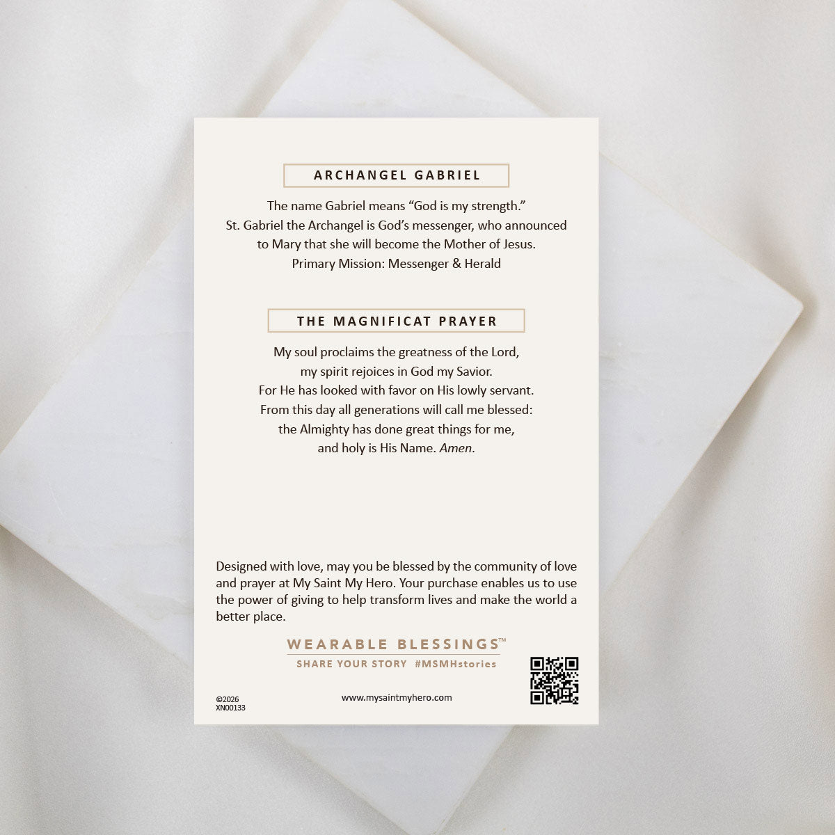 The back of a My Saint My Hero informational product card for the Archangel Gabriel Guardian Angel Necklace, resting on a white textured stone surface. The cream-colored card features the heading "ARCHANGEL GABRIEL" explaining his name and role as God's messenger, followed by the complete "THE MAGNIFICAT PRAYER". A QR code and the "WEARABLE BLESSINGS" branding are visible at the bottom.