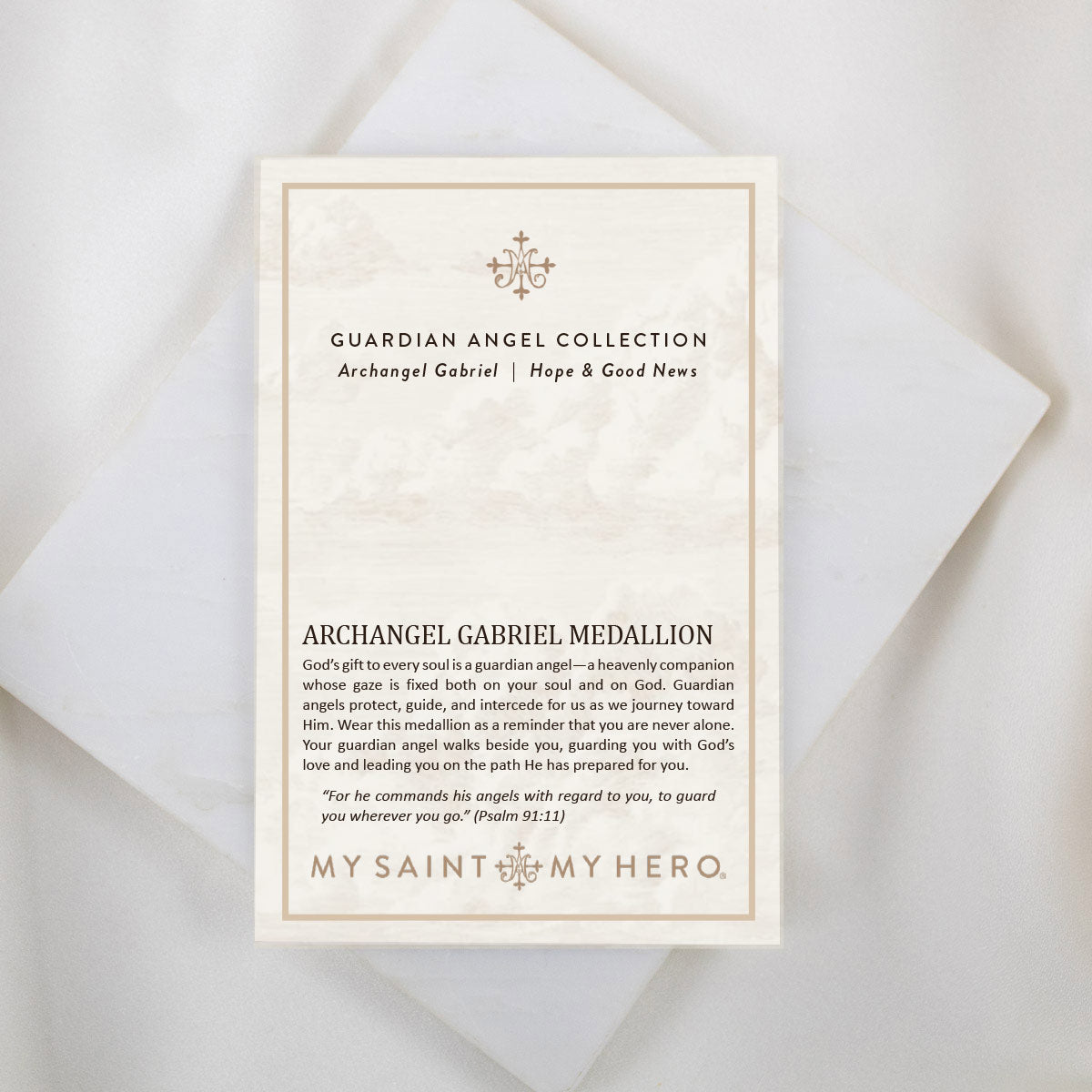 The front of a My Saint My Hero informational product card for the Archangel Gabriel Guardian Angel Necklace, resting on a white textured stone surface. The cream-colored card features the heading "GUARDIAN ANGEL COLLECTION" and "Archangel Gabriel | Hope & Good News" with a small decorative cross symbol at the top. The lower section is titled "ARCHANGEL GABRIEL MEDALLION" and features an inspirational paragraph about guardian angels alongside a quote from Psalm 91:11. The "MY SAINT MY HERO" brand logo is di