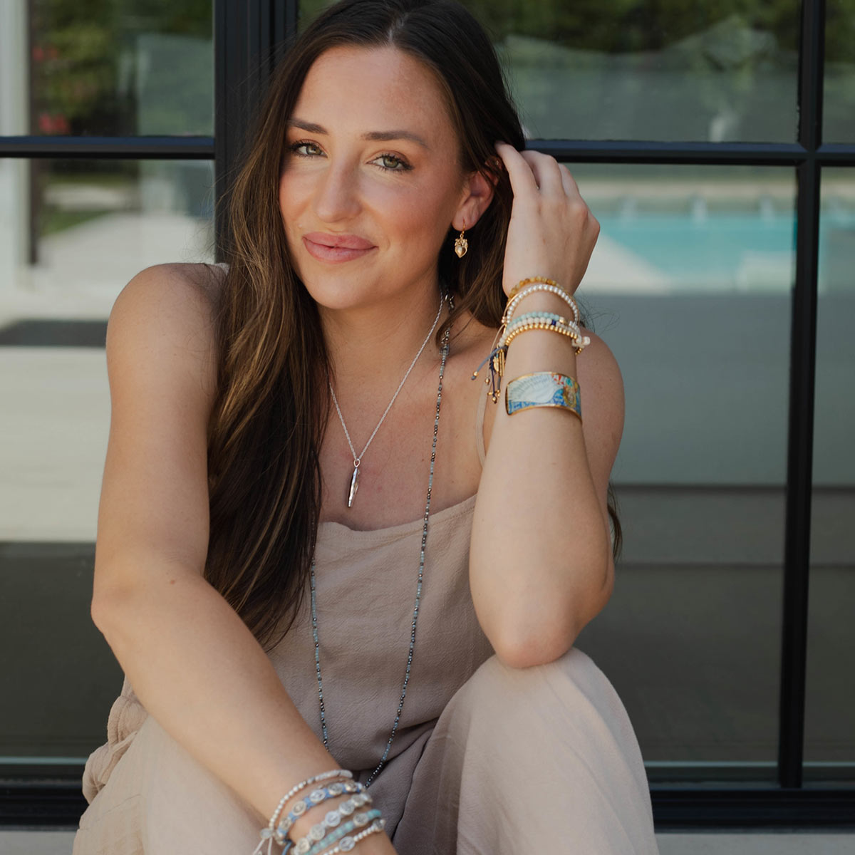 A woman wearing a silver beaded necklace, a silver feather pendant, and multiple bracelets sits in front of a glass window. She is wearing a beige sleeveless top and beige pants.