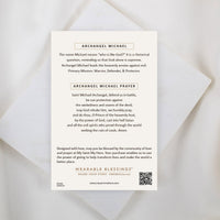 The back of a My Saint My Hero informational product card for the Archangel Michael Guardian Angel Necklace, resting on a white textured stone surface. The cream-colored card features the heading "ARCHANGEL MICHAEL" explaining his name and role as a warrior, defender, and protector, followed by the complete "ARCHANGEL MICHAEL PRAYER". A QR code and the "WEARABLE BLESSINGS" branding are visible at the bottom.