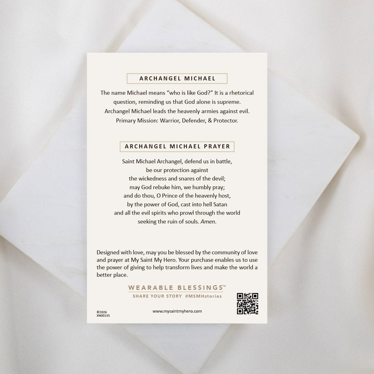 The back of a My Saint My Hero informational product card for the Archangel Michael Guardian Angel Necklace, resting on a white textured stone surface. The cream-colored card features the heading "ARCHANGEL MICHAEL" explaining his name and role as a warrior, defender, and protector, followed by the complete "ARCHANGEL MICHAEL PRAYER". A QR code and the "WEARABLE BLESSINGS" branding are visible at the bottom.