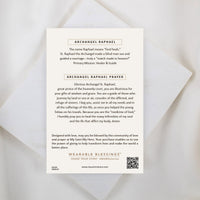 An inspirational product information card for the Archangel Raphael Guardian Angel Necklace, placed on a white marble background. The card reads "ARCHANGEL RAPHAEL" at the top, explaining that the name means "God heals," followed by the "ARCHANGEL RAPHAEL PRAYER" asking for guidance, healing, and protection. At the bottom, a "WEARABLE BLESSINGS" brand logo and a QR code are visible.
